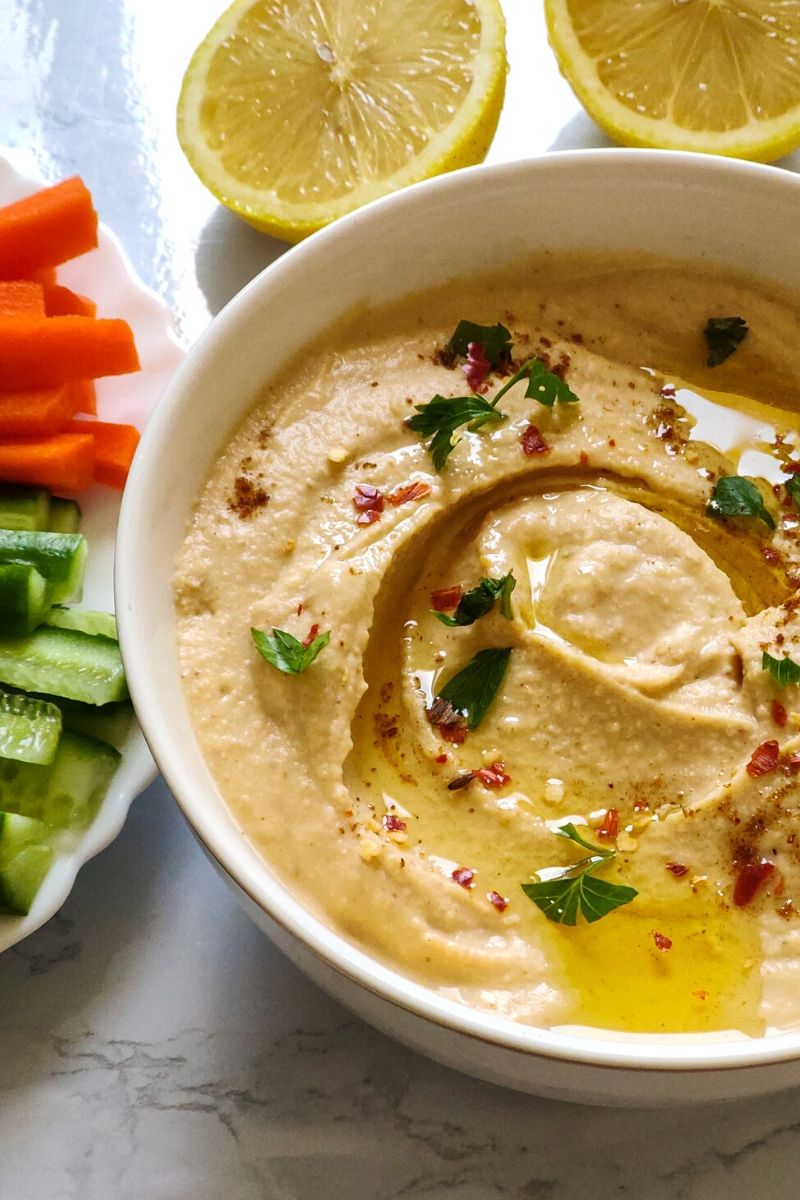 Hummus served in a white bowl with vegetable sticks and lemon wedges in the background