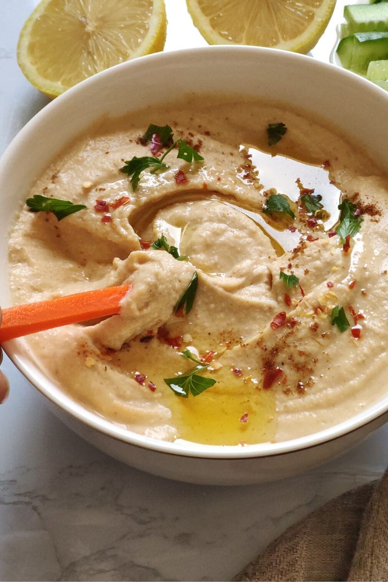 A carrot stick dipped in white bowl of hummus with two lemon wedges and more vegetable sticks in the background