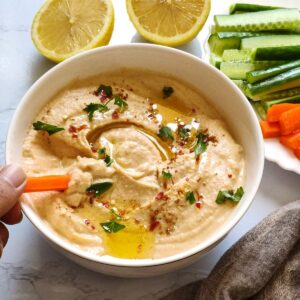 A carrot stick dipped in white bowl of hummus with two lemon wedges and more vegetable sticks in the background