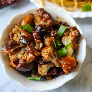 Kung pao cauliflower served in a white bowl with some noodles in the background