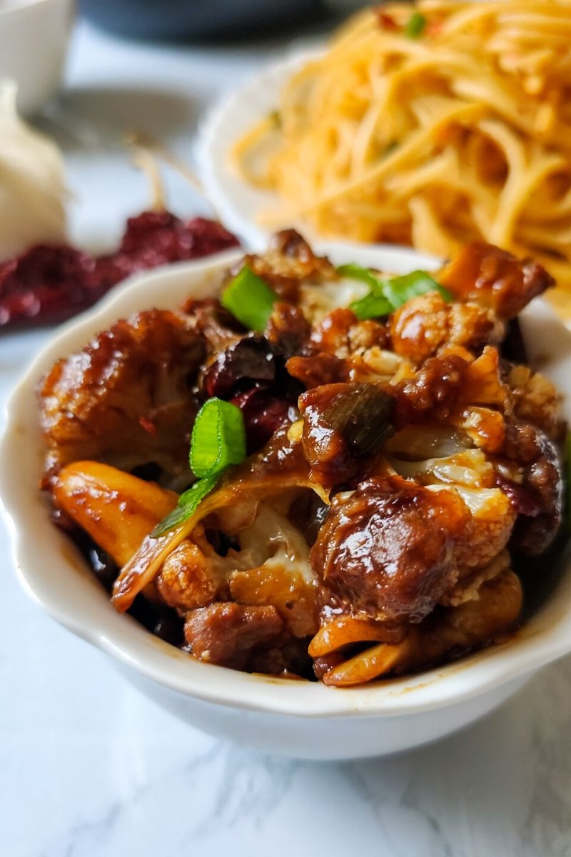 Kung pao cauliflower served in a white bowl with some noodles in the background