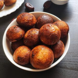 Sweet paniyaram served in a white bowl with some dates and paniyaram in the background