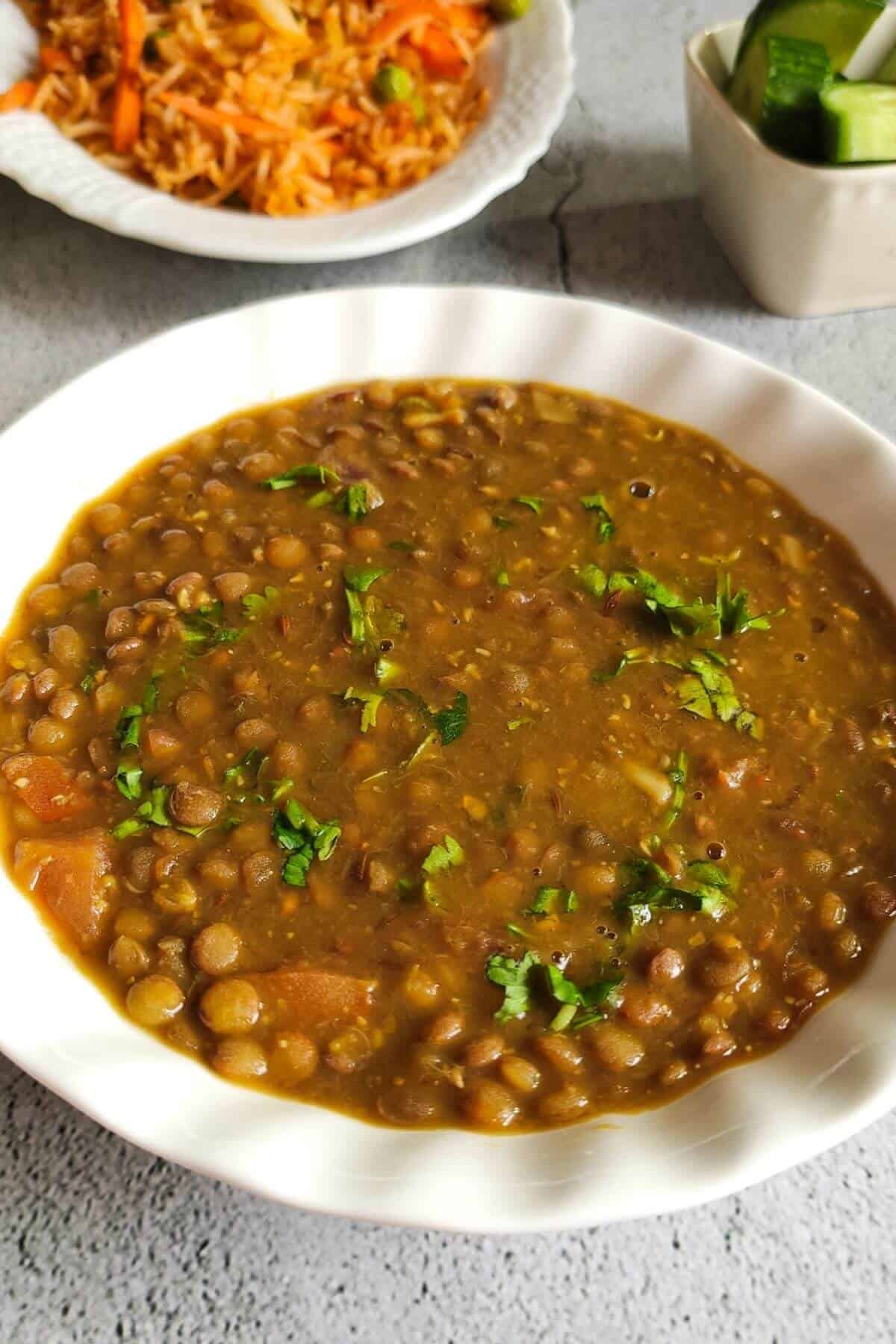 Brown lentil curry served in a bowl with rice and salad in the background
