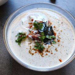 Aloo raita garnished with spices, cilantro, and curry leaves served in a glass bowl