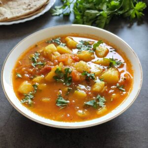 Indian potato curry served in a white bowl with chapati and cilantro in the background