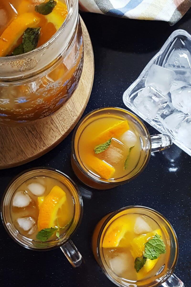 Orange iced tea served in three glasses and a pitcher with extra ice cubes in a bowl
