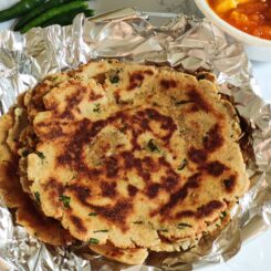 A pile of rajgira parathas kept on aluminium foil with some green chilies and curry in the background