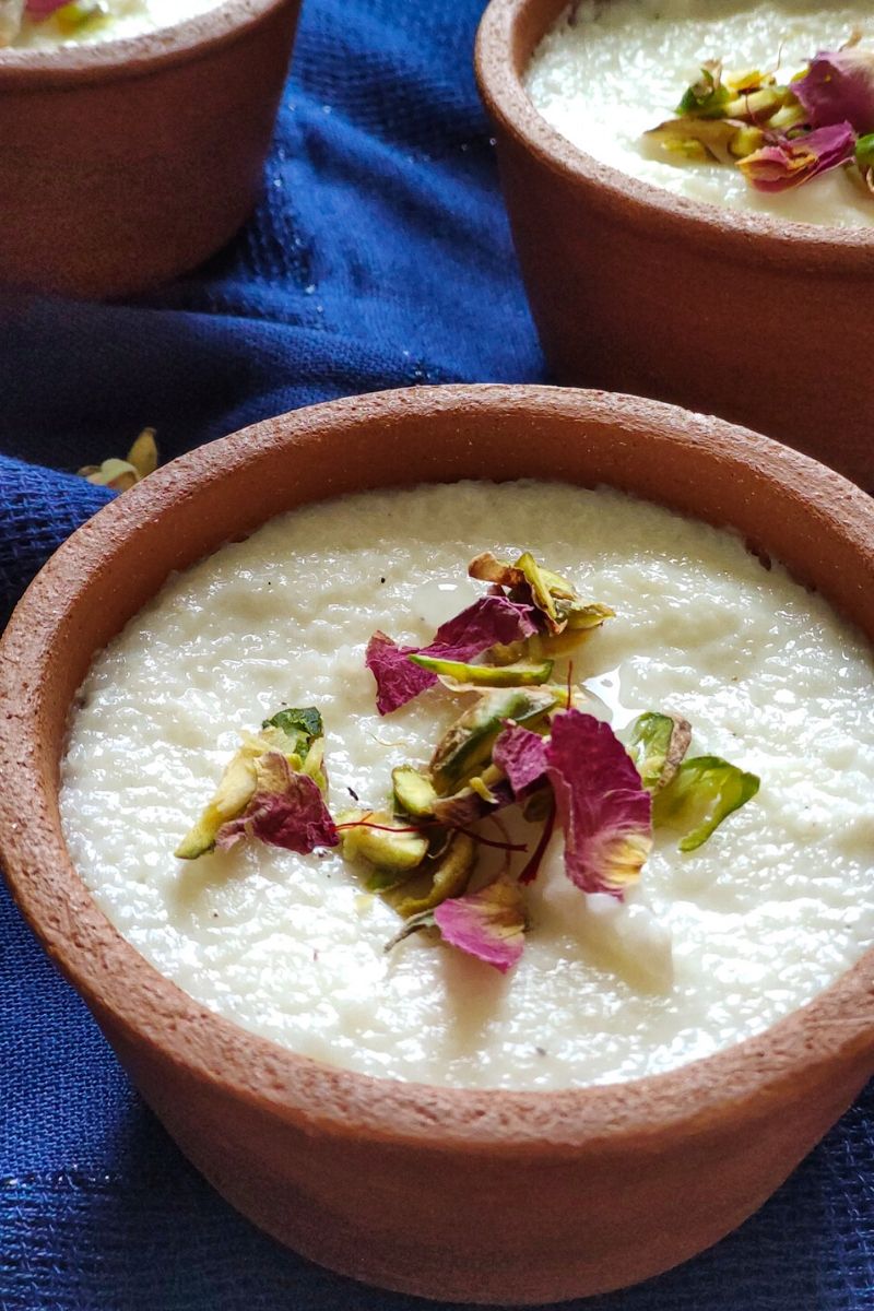 Indian rice pudding phirni garnished with dried rose petals, pistachio, and saffron served in two clay bowls kept on a blue table cloth