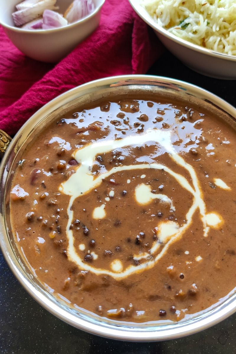 Dal makhani garnished with cream served in a bowl with some sliced onion and rice in the background