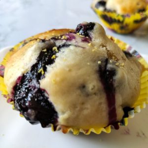 One vegan blueberry muffin in a yellow paper cup another muffin in the back ground