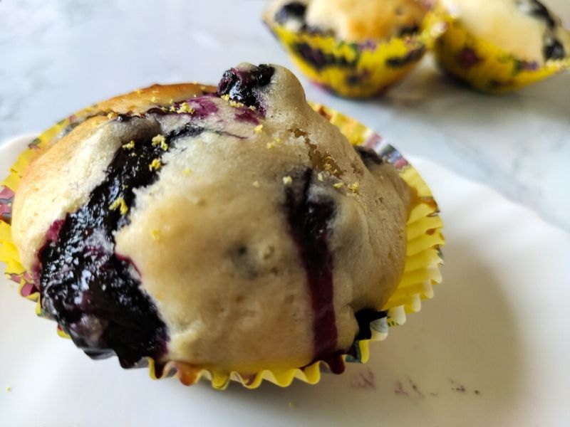 One vegan blueberry muffin in a yellow paper cup another muffin in the back ground