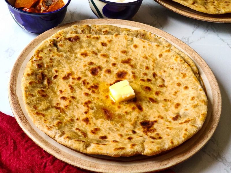 2 aloo parathas on top of each other topped with a butter cube served on a light brown plate with pickle and yogurt in the background