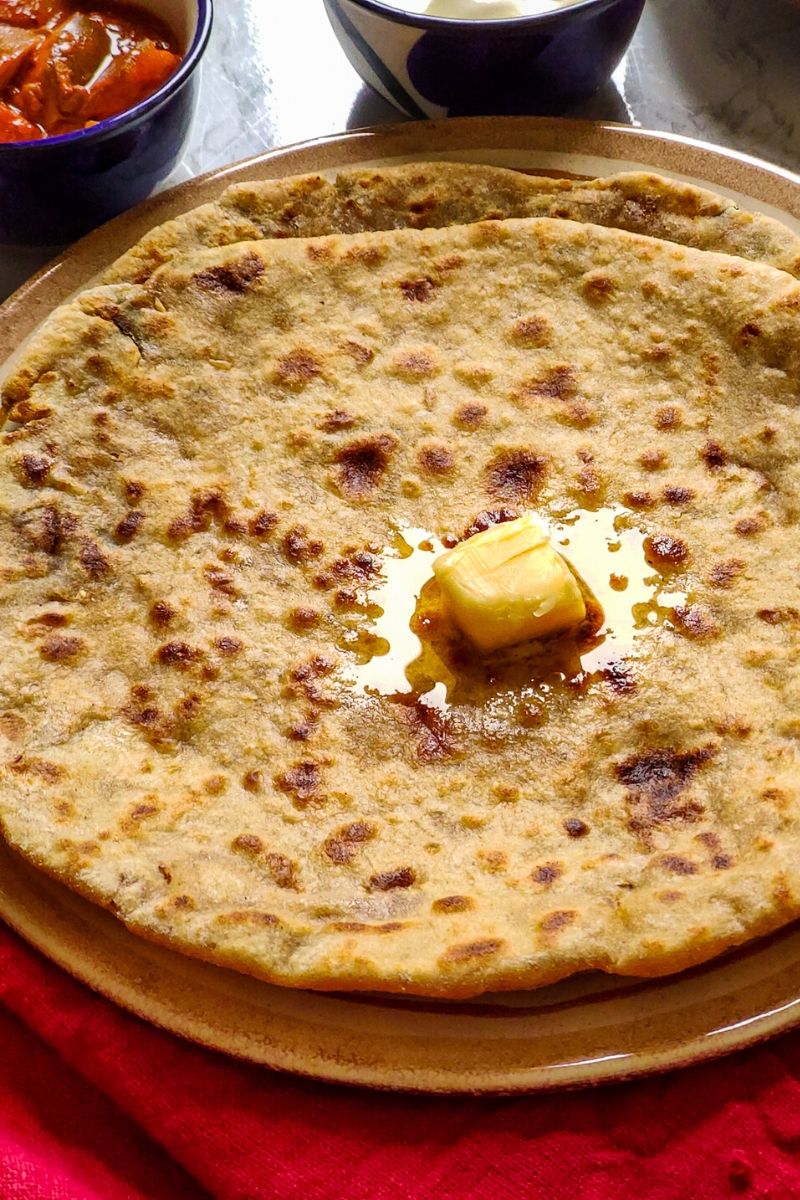 2 aloo parathas on top of each other topped with a butter cube served on a light brown plate with pickle and yogurt in the background