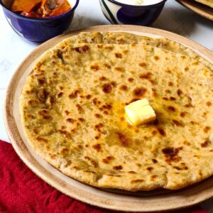 2 aloo parathas on top of each other topped with a butter cube served on a light brown plate with pickle and yogurt in the background