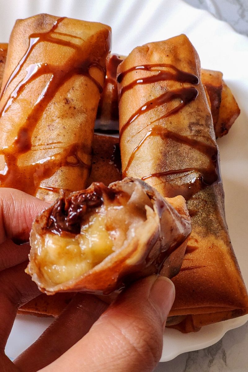 Half piece of chocolate banana spring rolls held in hand and more spring rolls served on a white plate in the background