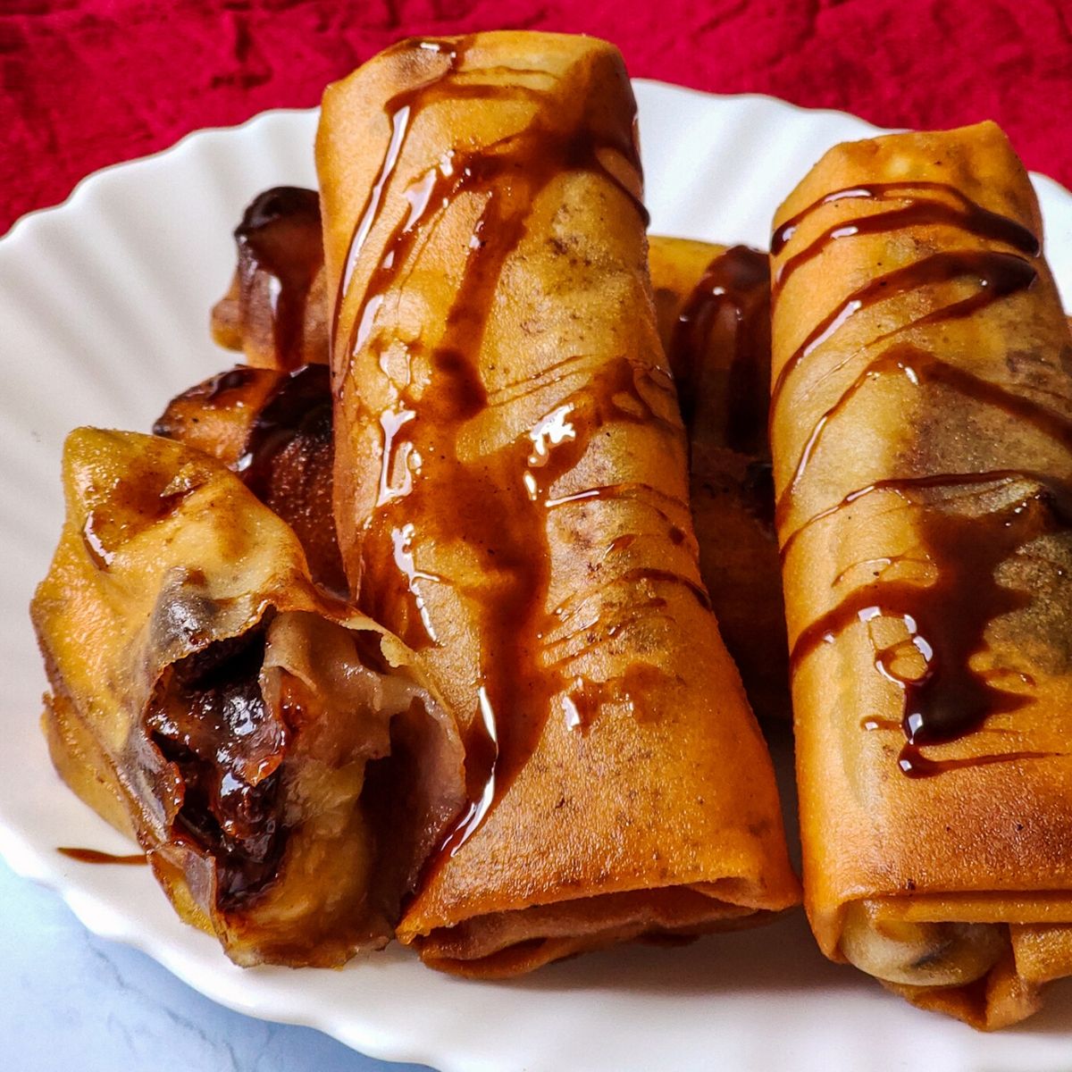 Chocolate Banana Spring rolls drizzled with chocolate sauce served on a white plate kept on a red table napkin