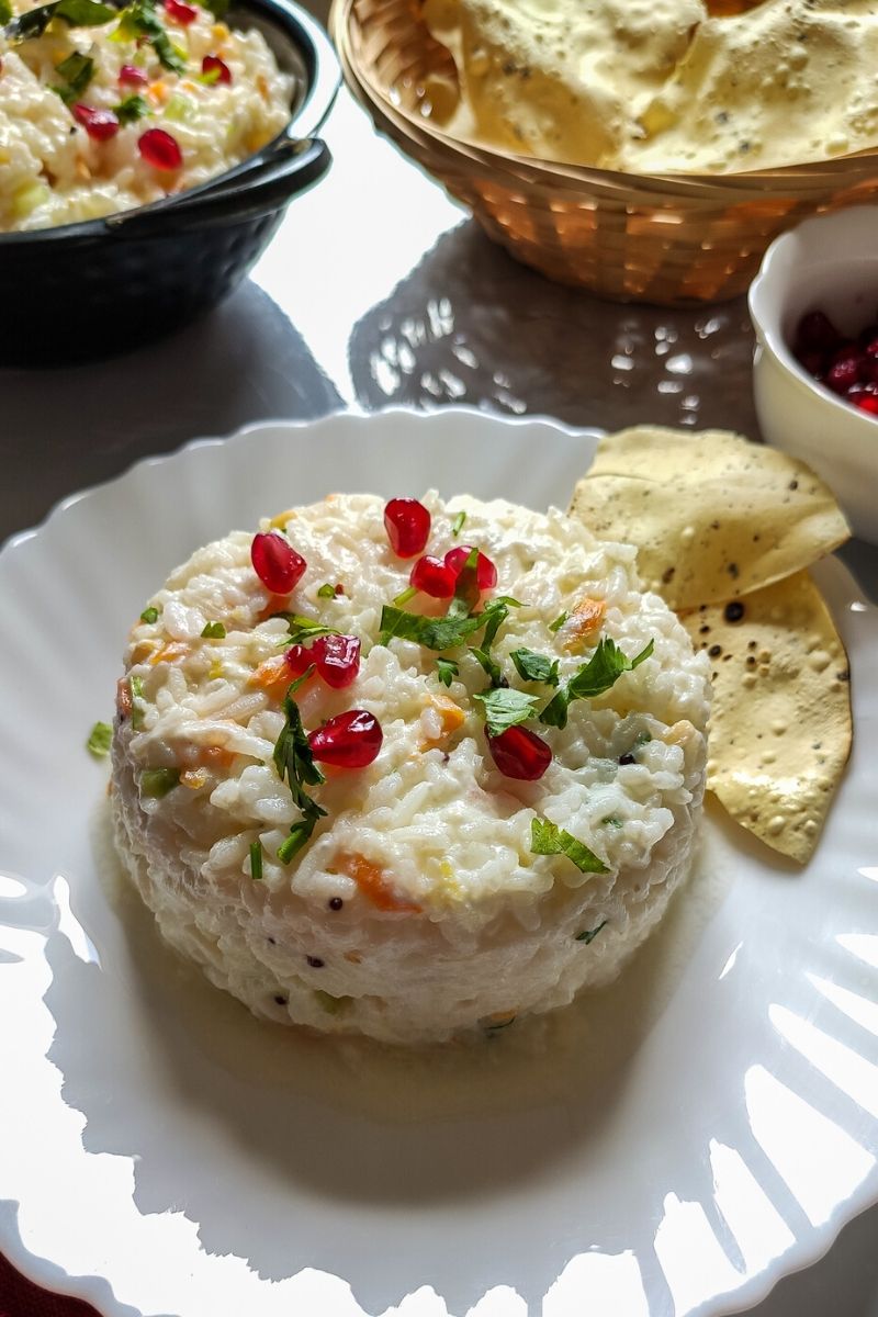 Curd rice garnished with pomegranate seeds served on a white plate with roasted popadums