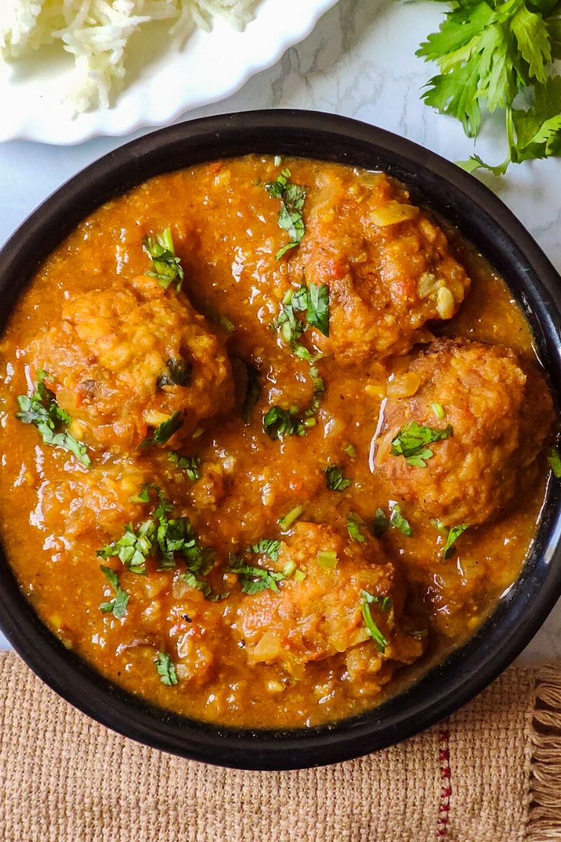 lauki ke kofte garnished with coriander leaves served in a black bowl with some rice and more coriander leaves in the background