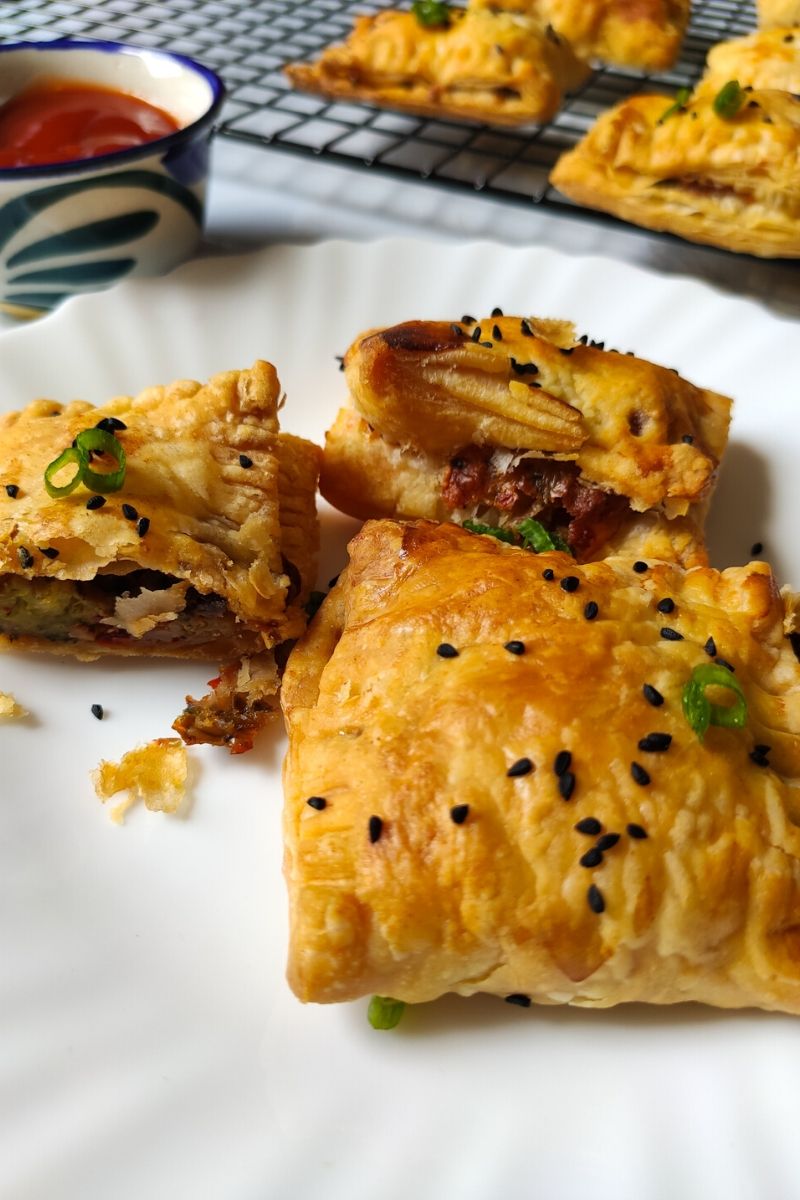 Veg pastry puffs kept on a white plate with more puffs and ketchup in the background