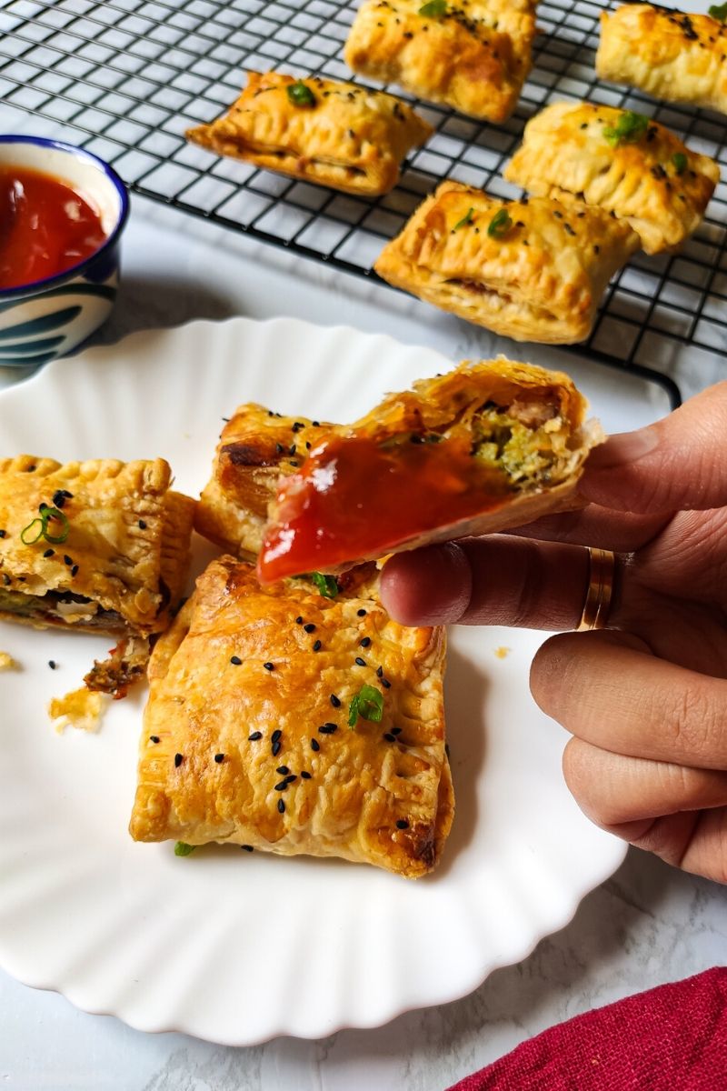 Vegetable puff dipped in sauce lifted above a plate of vegetable patties kept on a white plate