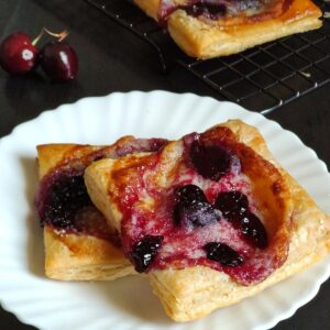 Two cherry cream cheese Danishes on a white plate with some cherries and another danish in the background