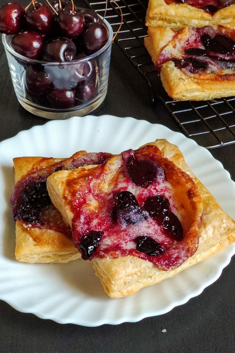 Two cherry cheese danishes on a white plate with a bowl of cherries and another danish in the background