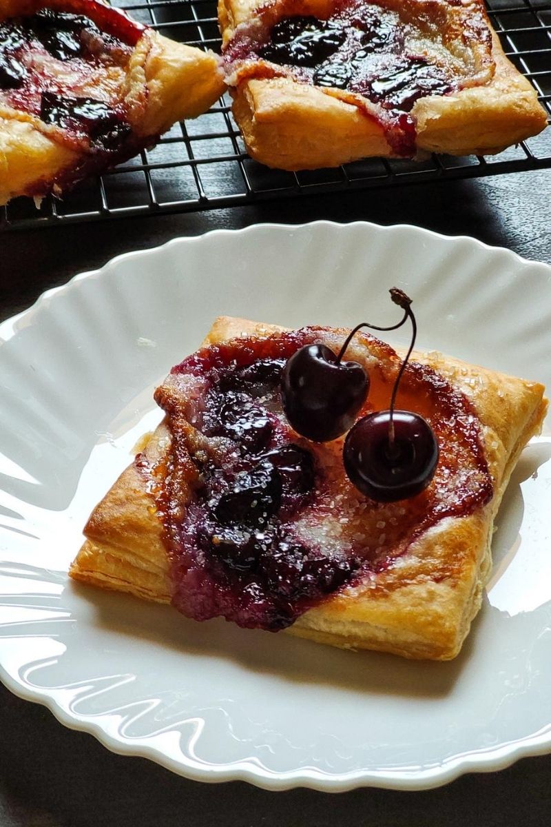 cherry cheese danish with cherries on top served on a white plate and more danishes in the background