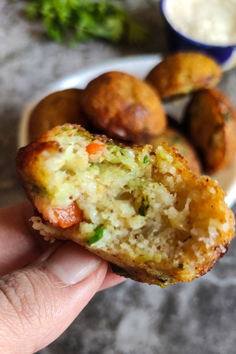 Half eaten vegetable paniyaram held over a bowl of paniyaram