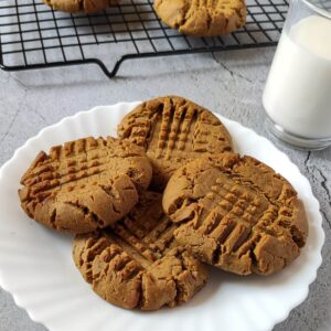 Peanut butter cookies on a white plate with a glass of milk and more cookies in the background.