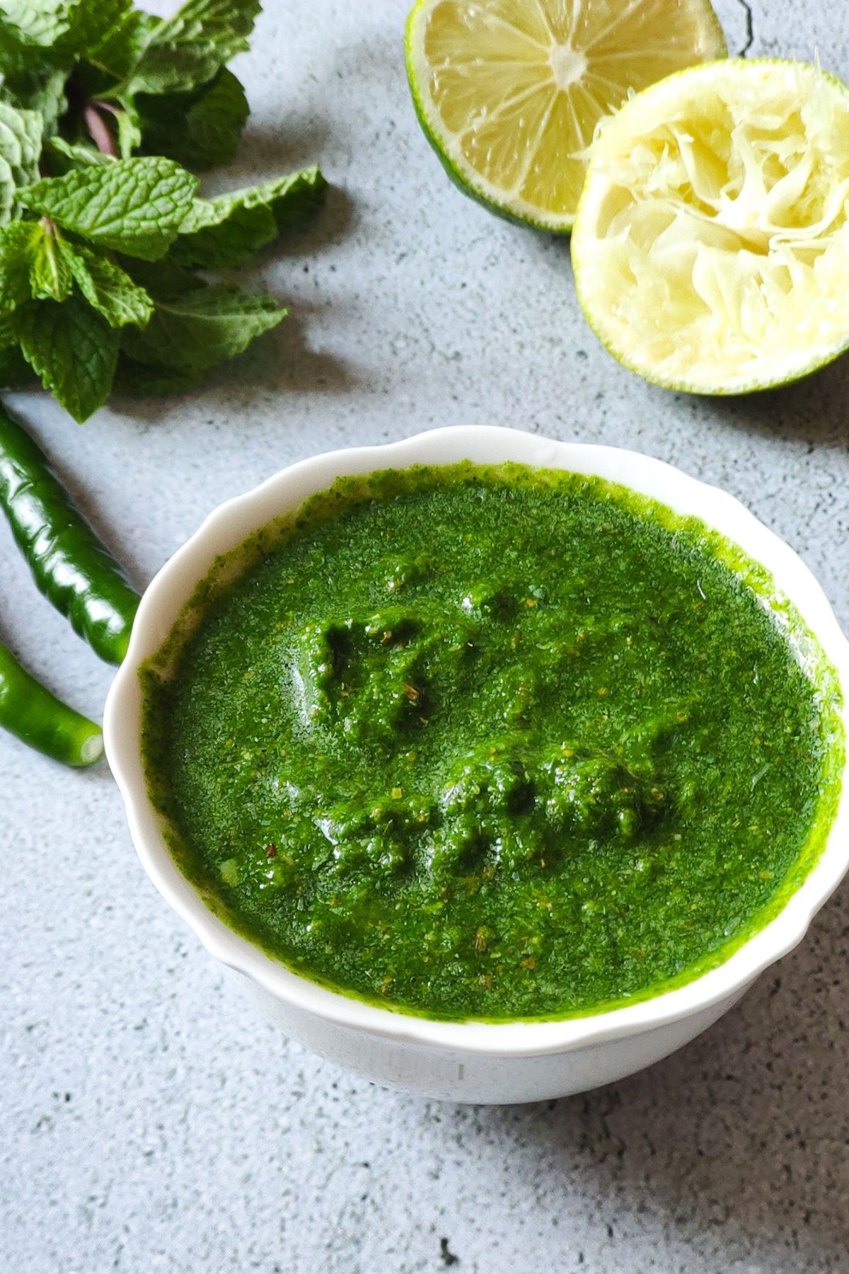 Green chutney in a white bowl with chilies, mint leaves, and lime in the background