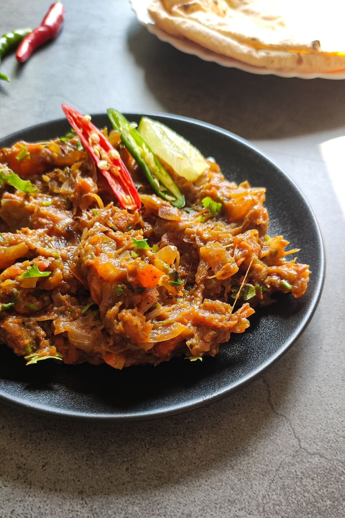 Baingan bharta in a black plate with chapati and chili peppers in the background