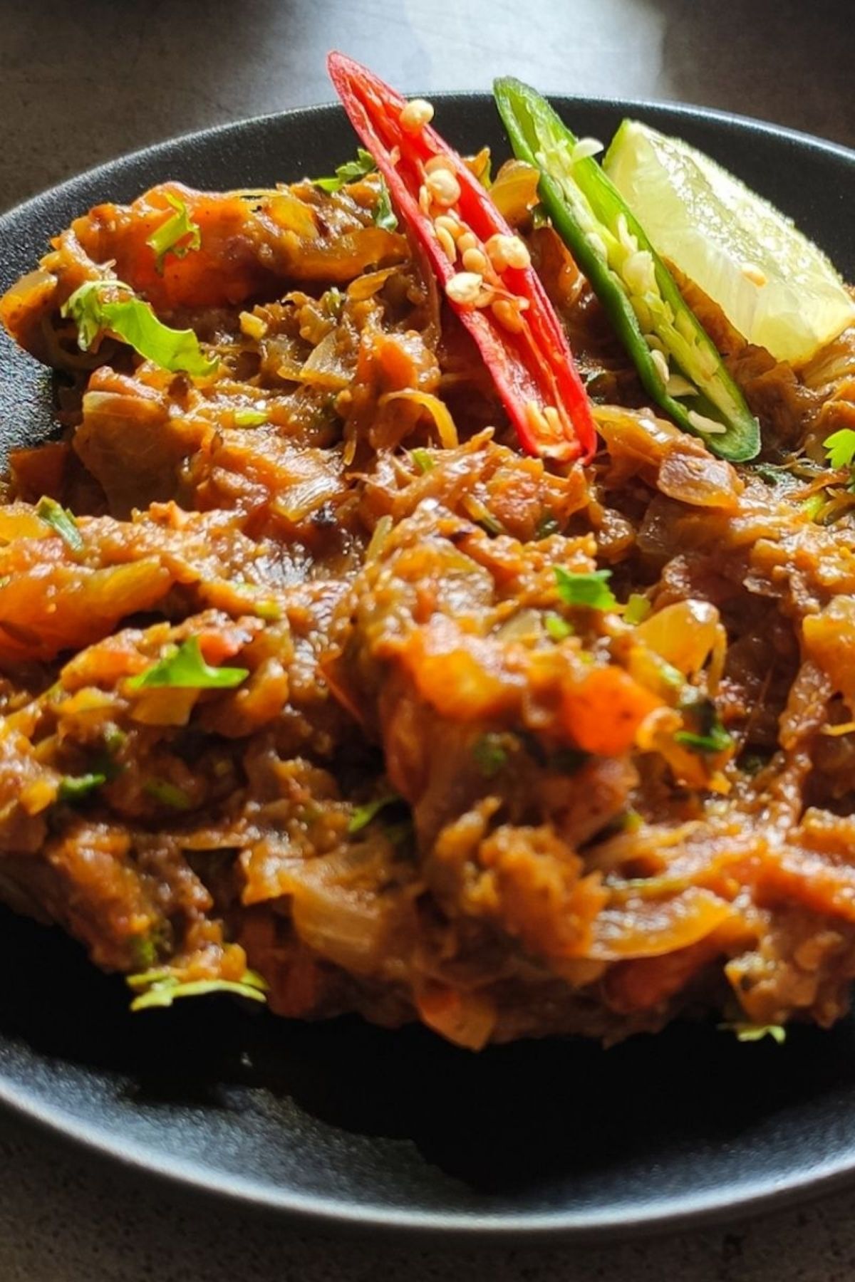 Baingan bharta on a black plate garnished with chili peppers and lemon wedge