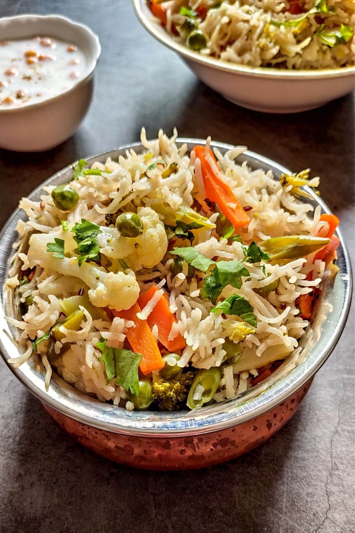 vegetable rice served in bowl with another bowl of rice and raita in the background