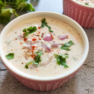 Onion raita served in a bowl with another bowl and herbs in the background
