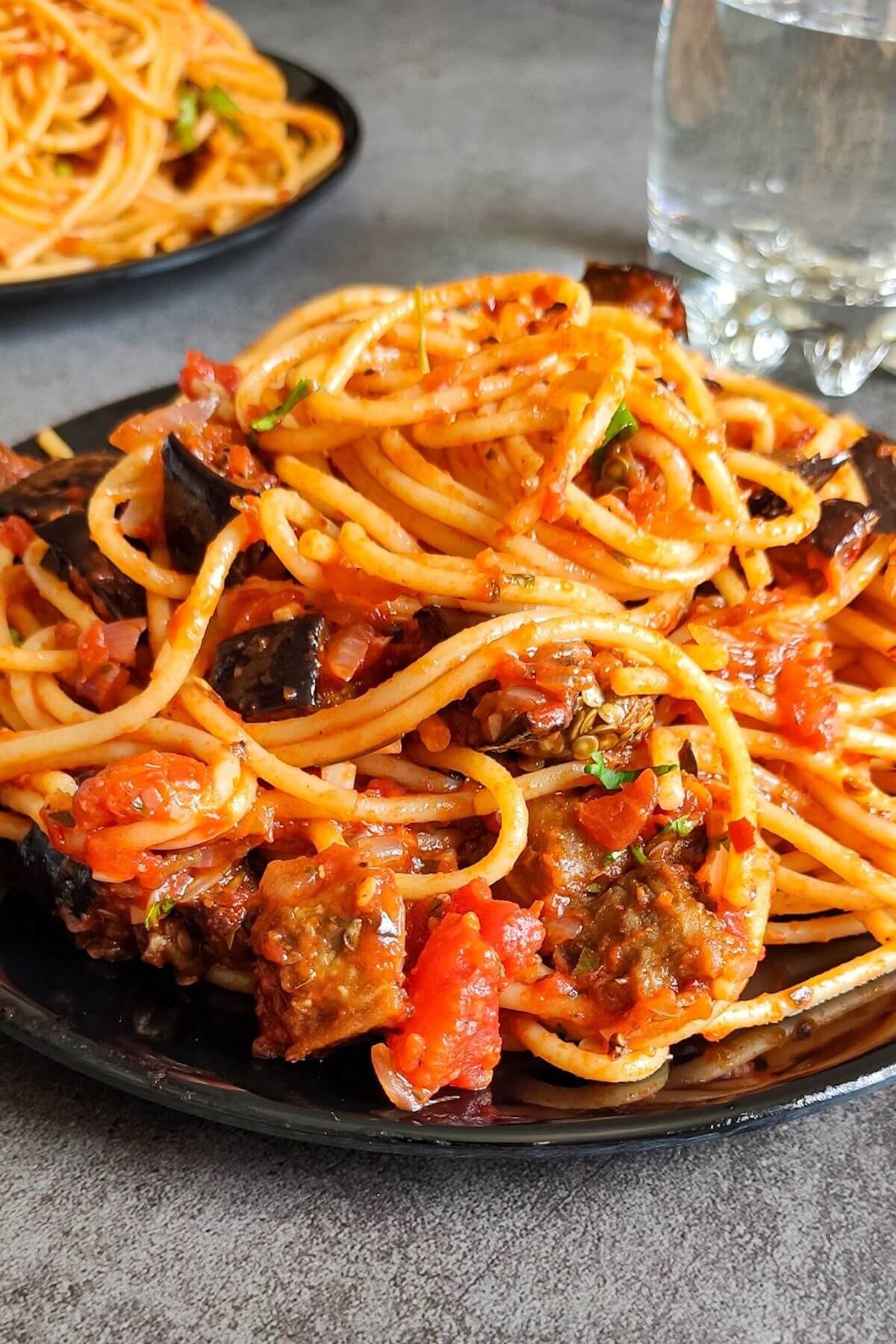 Eggplant pasta served on a black plate with a glass of water and more pasta in the background