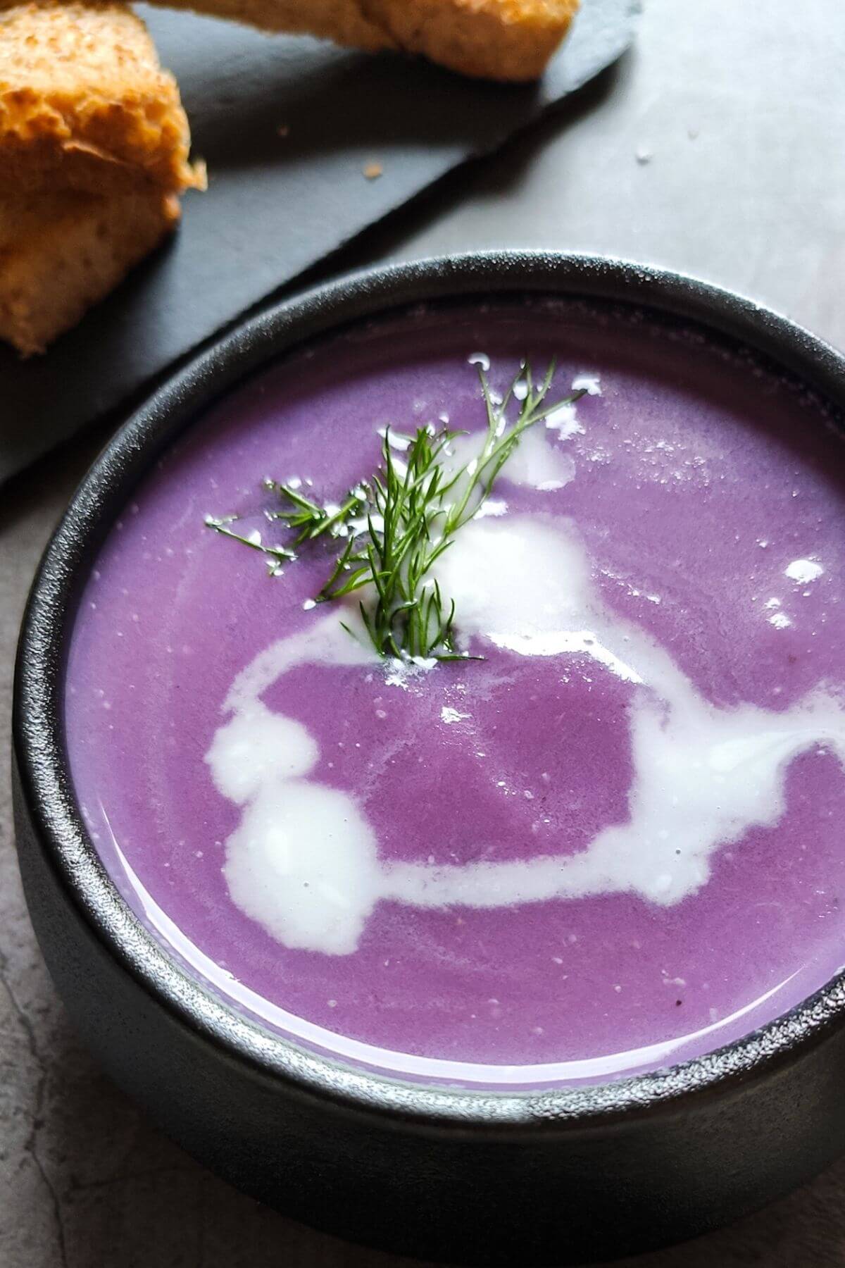 Red cabbage soup garnished with dill and almond milk served in a bowl with bread sticks in the background