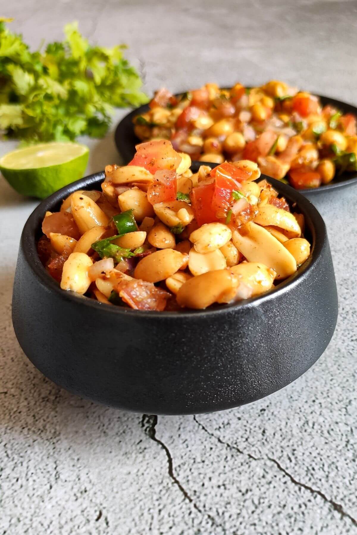 Quick roasted peanut chaat served in a black bowl and another plate of chaat, half a lime, and cilantro in the background