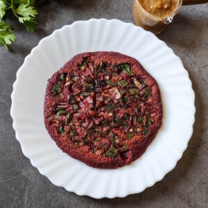Beetroot chilla served on a white plate with a bowl of dipping sauce and cilantro in the background