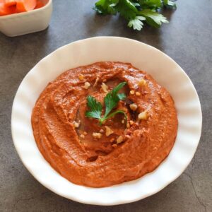 Muhammara dip garnished with chopped walnuts and parsley served in a white bowl.