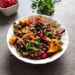 Winter lentil salad served in a white bowl with herbs and pomegranate arils in the background