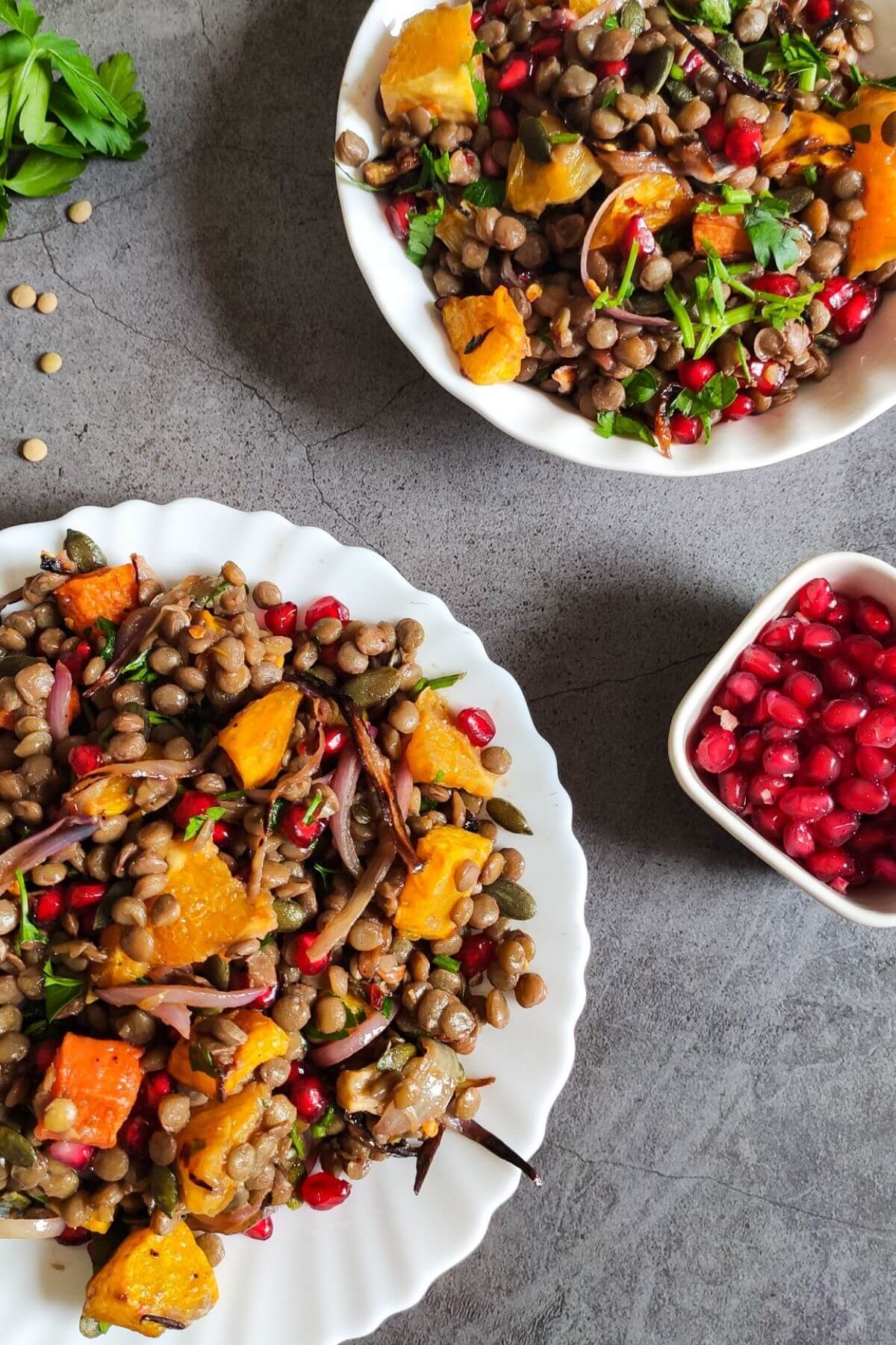 two plates of vegan green lentil salads with pomegranate arils in a bowl and fresh herbs on the side