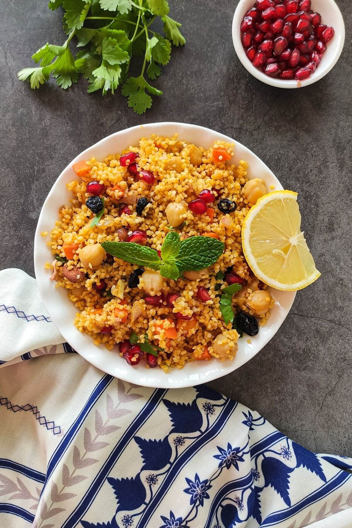 Moroccan couscous salad in a white bowl with fresh herbs and pomegranate arils in the background.