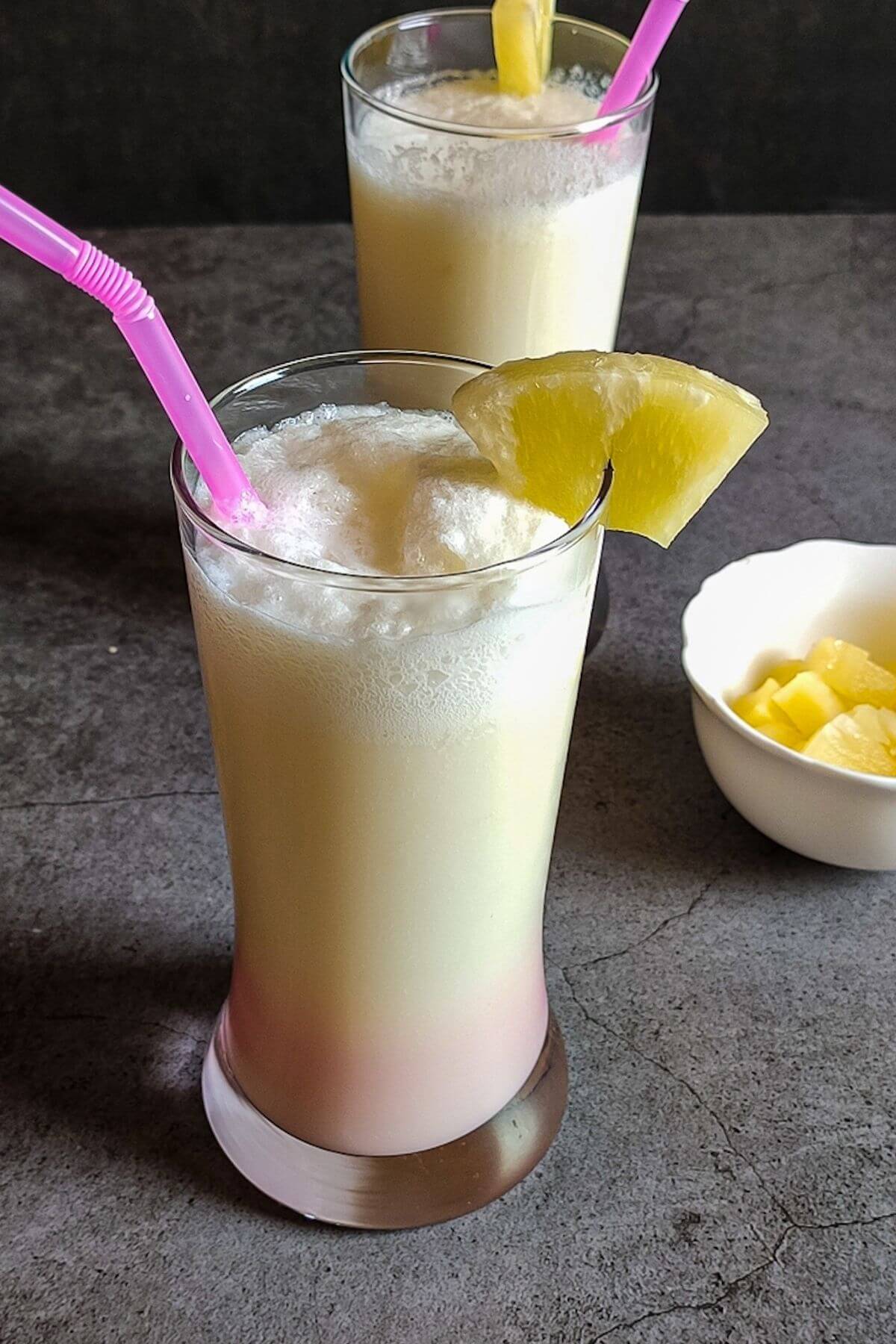 two glasses of virgin pina colada decorated with pineapple chunks and pink straw with a bowl of pineapple chunks on the side.