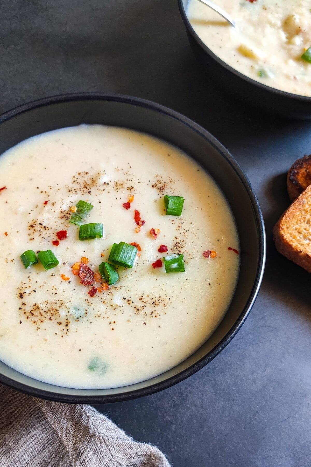 a bowl of potato soup garnished with spring onion greens and another bowl and toasts in the background.