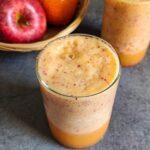 two glasses of apple orange smoothie with a basket of fruits in the background.