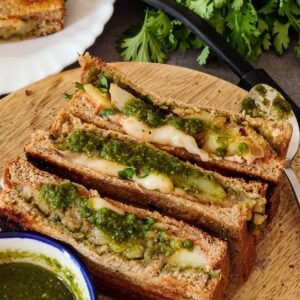 Three potato sandwiches with green chutney on a wooden board.
