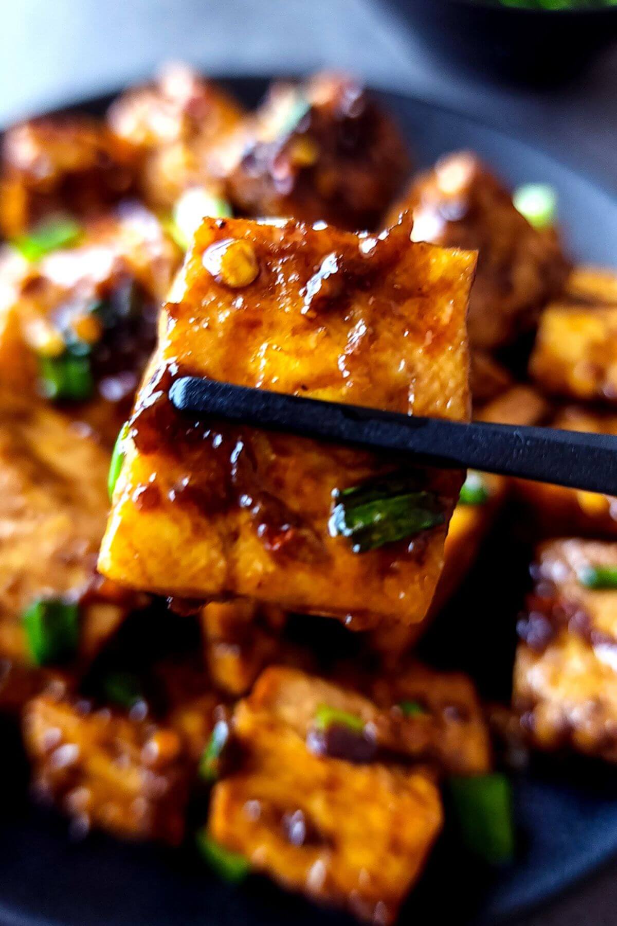 A tofu cube picked up with chopsticks over a plate of chili garlic tofu