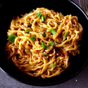 Peanut butter noodles garnished with green onions and chili flakes served in a black bowl.