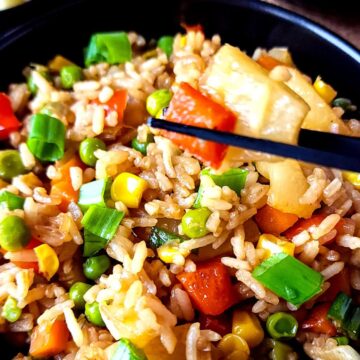 A piece of pineapple and red pepper lifted over a bowl of pineapple fried rice with a pair of chopsticks.