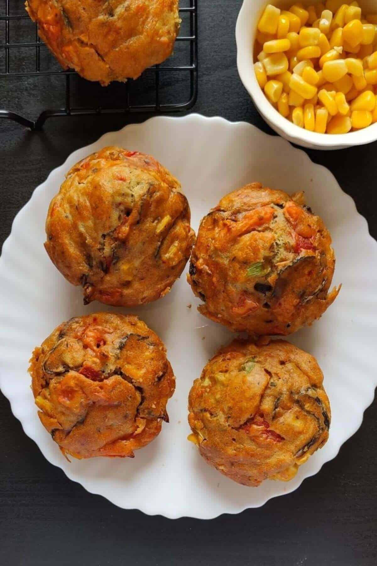 Four savoury muffins kept on a white plate with some corn kernels in the background
